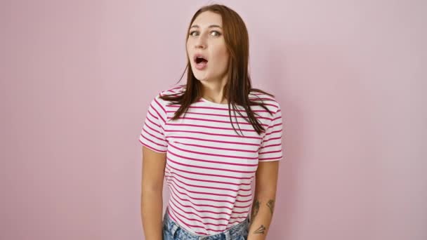 ピンクのバックグラウンドを覗くストライプTシャツの若いブルネットガールは 陽気でクレイジーな顔を作ります 楽しさと自信に満ちた表現 — ストック動画