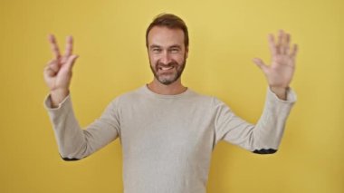 Neşeli İspanyol adam işaret ederken kendinden emin bir gülümseme sergiliyor, parmaklarıyla sekiz numarayı gösteriyor, izole edilmiş sarı bir duvara karşı duruyor..