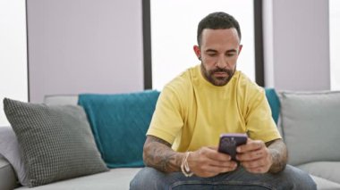 Modern oturma odasında akıllı telefon kullanan sakallı İspanyol bir adam.