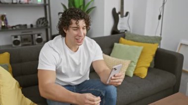 Modern bir oturma odasında anahtarlarını tutarken akıllı telefon kullanan kıvırcık saçlı neşeli bir genç adam..