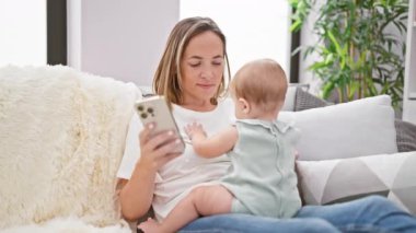 Sevgi dolu bir anne ve mutlu bir kız evdeki kanepede oturarak serin ve rahat bir anı paylaşıyorlar. Kendine güvenen bir kadın gülümseyen bebeğiyle meşgul ve akıllı telefonuna bir mesaj yazıyor..