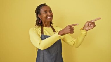 Neşeli orta yaşlı Afro-Amerikalı önlüklü kadın, izole edilmiş sarı arka planda, kameraya ışıldarken kendine güvenerek duruyor..