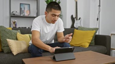 Evde tablet kullanan odaklanmış İspanyol bir adam rahat bir oturma odasında modern teknolojiyi sergiliyor..
