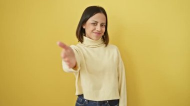 Sarı süveterli neşeli genç İspanyol kadın, sarı bir arka planda yalnız duruyor, gülümsüyor ve dostça bir selamlaşma olarak başarılı bir tokalaşma teklif ediyor.