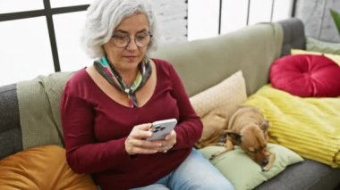 Gri saçlı kadın, evde kanepede uyuyan köpekle akıllı telefon kullanıyor.