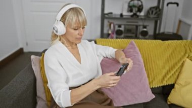 Orta yaşlı bir kadın, renkli yastıkları ve modern dekorları olan rahat bir oturma odasında kulaklıkla ve akıllı telefonuyla rahatlar.
