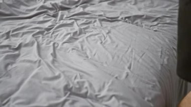 Kimliği belirsiz bir adam, kırışık bir çarşafın üzerinde oturuyor ve gri bir yatak odasına giriyor..