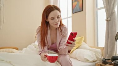 Pijamalı kızıl saçlı kadın akıllı telefon kullanıyor ve rahat bir yatak odasında kahve içiyor..