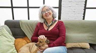 Gri saçlı bir kadın, rahat bir oturma odasında köpeğiyle rahat bir kanepede vakit geçirmek ister..