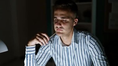 Çizgili tişörtlü düşünceli İspanyol adam loş ofis alanında kalem tutuyor.