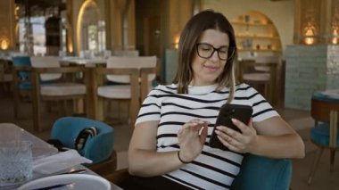 Akıllı telefon kullanan gülümseyen bir kadın şık bir restoranda selfie çeker, modern bir yemek ortamında bağlantı ve eğlence tasvir eder..