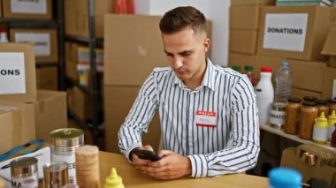 Alex adında genç bir adam bağış merkezinde akıllı telefon kullanırken gülümsüyor. Arka planda çeşitli yiyecek ve kutular var..