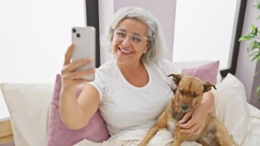 Gülümseyen gri saçlı kadın, köpeğiyle sıcak yatak odasında selfie çekiyor..
