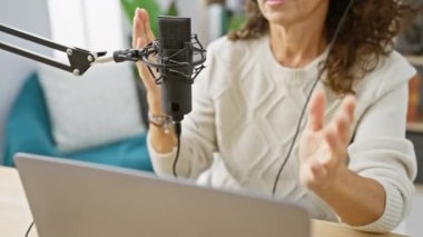 Orta yaşlı İspanyol bir kadın, stüdyo masasında dizüstü bilgisayarla mikrofona konuşan bir podcast kaydediyor..