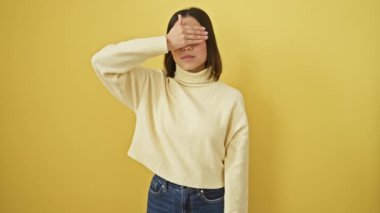 Şok olmuş ve utangaç, kazaklı güzel bir kadın parmaklarından süzülüyor, yüzünü örtüyor, elleri ürkek bir sürpriz sergiliyor! Sarı izole edilmiş arkaplanda göz teması