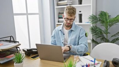 Mavi gözlü ve sakallı yakışıklı genç adam modern bir ofiste çalışırken sandviç yiyor.