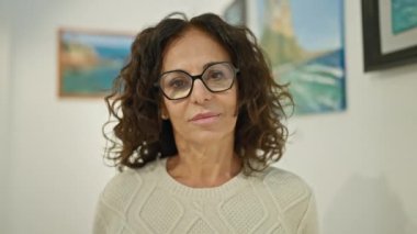 Gözlüklü, süveterli neşeli orta yaşlı bir kadın bir galeride resimlerle poz veriyor..