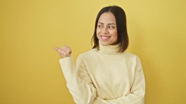 Süveter giyen neşeli genç İspanyol kadın başparmağını kaldırıyor, gülümsüyor, yan yana işaret ediyor. Sarı izole edilmiş arka planda ürün reklamı.