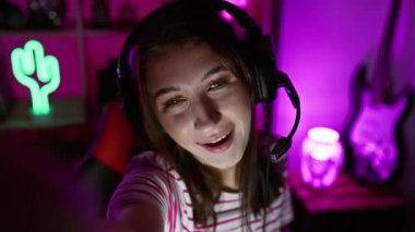 Kulaklıklı genç bir kadın oyun odasında selfie çekiyor. Arka planda neon ışıkları ve gitarı var. Modern, teknoloji meraklısı bir atmosferi aktarıyor..