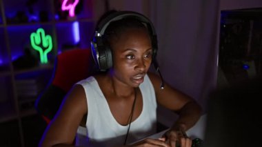 Kıvırcık saçlı Afrikalı Amerikalı bir kadın geceleri oyun odasında kulaklıklarını çıkarıyor..