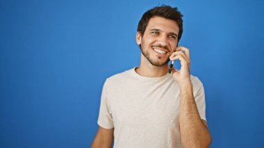 Sakallı, neşeli, genç, İspanyol bir adam bej bir tişört giyiyor ve cep telefonuyla dışarıda mavi bir duvara karşı konuşuyor..