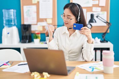 Ofiste çalışan genç Çinli bir kadın online alışveriş yapıyor. Başparmağıyla yan tarafa işaret ediyor. Ağzı açık gülümsüyor. 