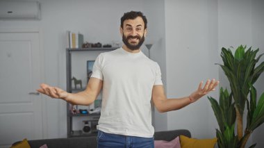 Beyaz tişörtlü, gülümseyen sakallı bir adam modern bir oturma odasında kollarını açmış, hoş bir atmosfer yayıyor..