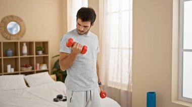 Genç bir İspanyol adam, iyi aydınlatılmış bir yatak odasında dambıllarıyla egzersiz yapıyor, sağlık ve rutin bir hissi uyandırıyor..