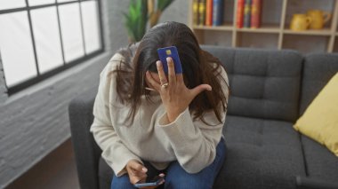 Stresli kadın kredi kartından alnına kadar akıllı telefonlu bir evde koltukta oturuyor..