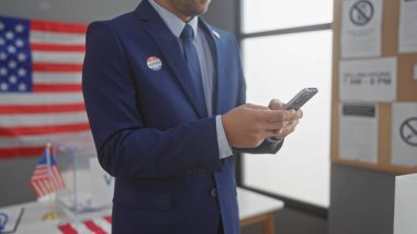 Takım elbiseli, sakallı genç bir adam Amerikan seçim merkezinde telefonunu kontrol ediyor. Bayrak ve oy kullanma kabinleriyle..