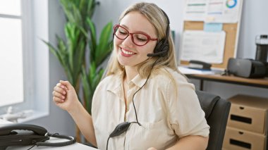 Ofiste kulaklık takan, bilgisayar ve dosyaları olan neşeli genç bir kadın profesyonel müşteri hizmetlerini somutlaştırıyor..