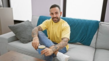 Rahat dövmeli bir adam, rahat bir oturma odasında, mavi bir kanepede kahveden zevk alır, modern ev konforunu somutlaştırır..