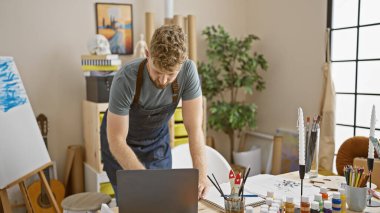 Etrafı resimler ve yaratıcı malzemelerle çevrili, parlak bir sanat stüdyosunda dizüstü bilgisayarda çalışan sakallı genç bir adam..