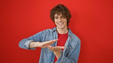 Kıvırcık saçlı yakışıklı genç adam kırmızı duvara karşı bir şey tutuyor gibi yapıyor, sıradan bir kot giysisi sergiliyor..