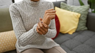 Kanepede oturan kıvırcık saçlı, rahat bir oturma odasında bileğini tutan yetişkin bir kadın..