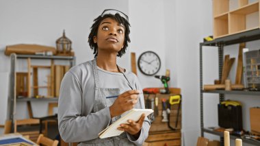 Düşünceli, Afro-Amerikalı, rastalı bir kadın, marangozluk stüdyosunda gri bir önlük giyiyor, notlar alıyor ve bir proje planlıyor..