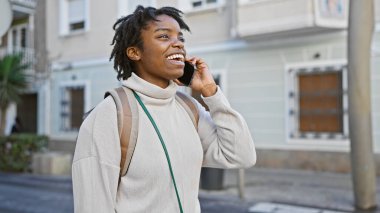 Rasta saçlı genç siyahi bir kadın şehir caddesinde akıllı bir telefonla konuşuyor.