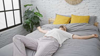 Sakallı genç beyaz bir adam, modern bir yatak odasındaki bir yatağa yayılmış, rahatlama ya da yorgunluğu ifade ediyor..