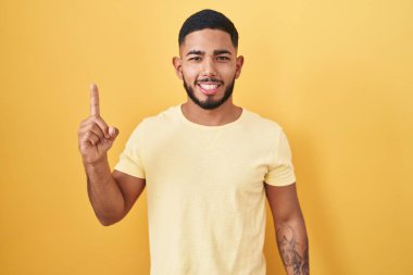 Sarı arka planda duran İspanyol genç adam bir numarayı gösterip işaret ederken kendinden emin ve mutlu bir şekilde gülümsüyor.. 