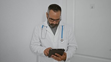 Beyaz laboratuvar önlüğü giyen sakallı ve gözlüklü olgun bir adam klinik koridorunda bir tablet okuyor..