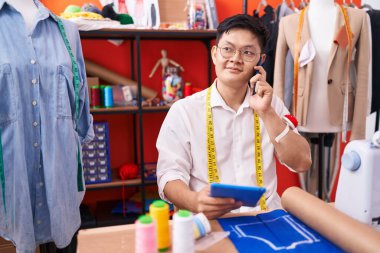Young chinese man tailor talking on smartphone using touchpad at atelier