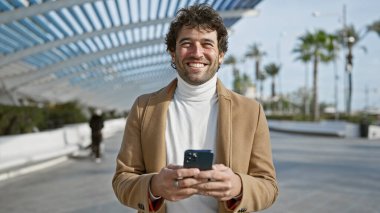 A handsome young hispanic man smiles while using a smartphone outdoors in a sunny, palm-lined urban park. clipart