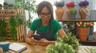 Yeşil önlüklü ve gözlüklü bitkilerle dolu bir çiçekçide akıllı telefon kullanan Afrikalı Amerikalı kadın çiçekçi..