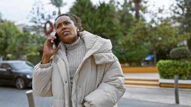 Endişeli genç bir kadın şehir caddesinde, üzerinde kışlık bir palto ve şehir yeşillikleriyle dikilirken telefonuyla konuşur..