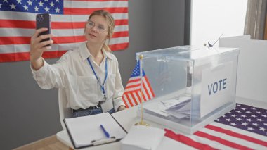 Genç bir kadın oy sandığı ve ön planda Amerikan bayrağı olan bir Amerikan oy merkezinde selfie çekiyor.