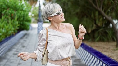 Yeşil bir parkta güneşli bir günün tadını çıkaran, güneş gözlüğü takan ve şık bir kıyafet giyen zarif, orta yaşlı bir kadın..