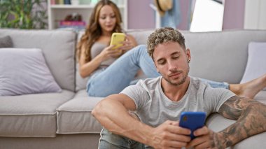 Dövmeli bir adam ve bir kadın, modern bir oturma odasındaki gri bir koltukta akıllı telefonlarıyla meşgul oluyorlar..