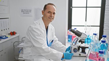 Mutlu orta yaşlı adam, kendine güvenen İspanyol bilim adamı, tıbbi laboratuarda mikroskop altında numune incelerken sırıtıyor.