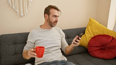 Sakallı, orta yaşlı bir adam evinde elinde akıllı bir telefon ve rahat bir oturma odasında kırmızı bir kupa tutarak rahatlıyor..