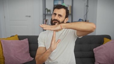 Orta yaşlı sakallı bir adam modern bir oturma odasında mola işareti yapıyor.